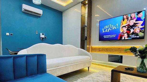 Een moderne woonkamer met een witte bank, een blauwe slaapbank en een flatscreen televisie waarop een programma draait.