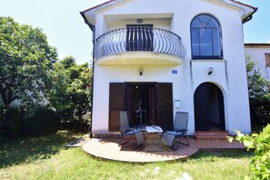 Un edificio de dos pisos con un balcón, un patio con sillas y una puerta que da a un espacio interior.