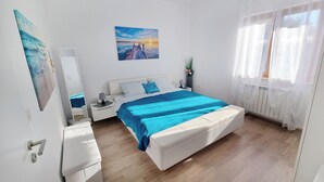 Ein Schlafzimmer mit einem Bett, einem Nachttisch, einer Kommode, einem Fenster mit Vorhängen und einem Wandbild.