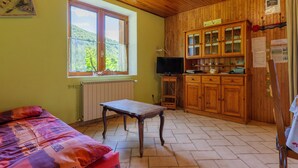 A room with a bed, a table, a TV, wooden cabinets, and a window with a view of greenery. - Gîte "À l’ombre du Tilleul": Mountain View, Private Terrace & Wi-Fi (Montpezat-sous-Bauzon)