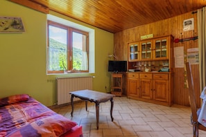 A room with a bed, a table, a TV, wooden cabinets, and a window with a view of greenery. - Gîte "À l’ombre du Tilleul": Mountain View, Private Terrace & Wi-Fi (Montpezat-sous-Bauzon)