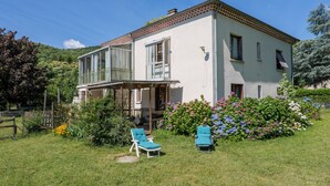 A two-story house with a balcony, a covered porch, and a garden with flowers and shrubs. - Gîte "À l’ombre du Tilleul": Mountain View, Private Terrace & Wi-Fi (Montpezat-sous-Bauzon)