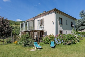 A two-story house with a balcony, a covered porch, and a garden with flowers and shrubs. - Gîte "À l’ombre du Tilleul": Mountain View, Private Terrace & Wi-Fi (Montpezat-sous-Bauzon)