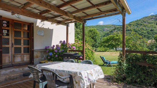 A wooden deck with a table and chairs, a covered patio, and a view of a green hillside. - Gîte "À l’ombre du Tilleul": Mountain View, Private Terrace & Wi-Fi (Montpezat-sous-Bauzon)
