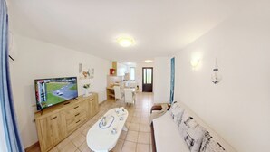 Een moderne woonkamer met een flatscreen-tv, een eethoek met een witte tafel en stoelen, een keuken op de achtergrond en een gang naar een deur.