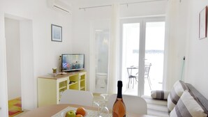 Ein Wohnzimmer mit Fernseher, einem Esstisch mit Früchten und Weingläsern, einer Couch und Meerblick durch die Tür.