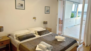 Ein Schlafzimmer mit Bett, Kissen, einem Nachttisch, einer Lampe und Blick durch eine Glastür auf einen Außenbereich.