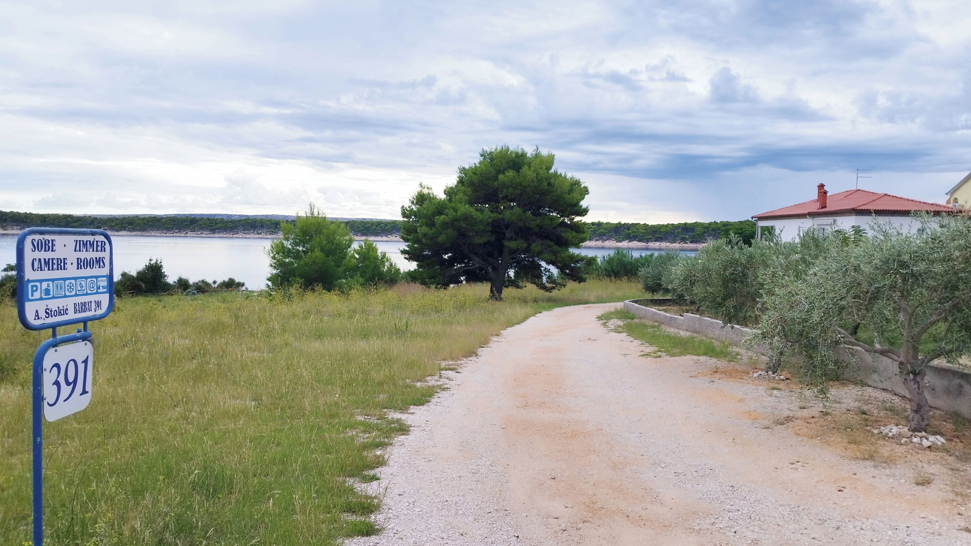 Un panneau indiquant les différents types d’hébergement, un chemin de terre, un arbre et une étendue d’eau en arrière-plan.