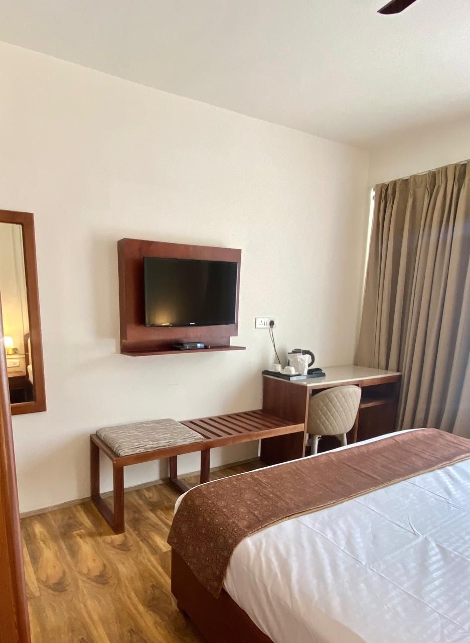 A hotel room with a bed, a desk with a chair, a television mounted on the wall, and a window with curtains.