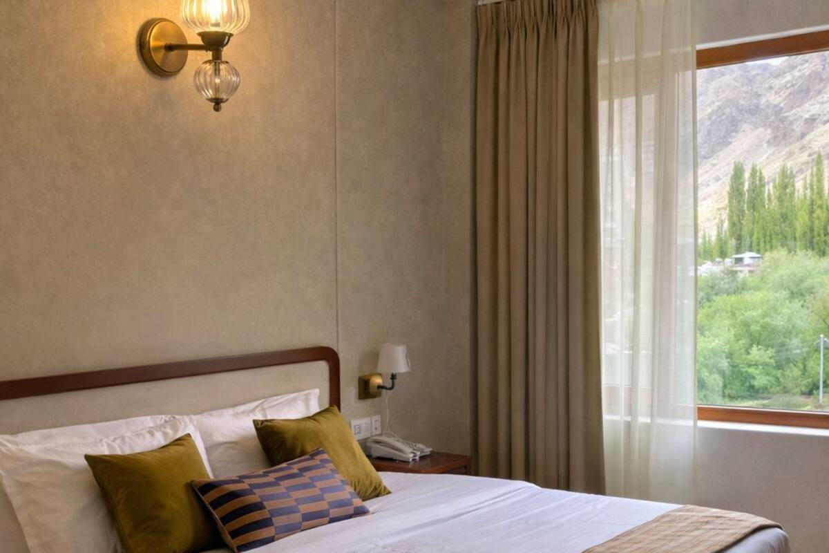 A bedroom with a bed, pillows, a bedside table, a lamp, and a window with curtains.