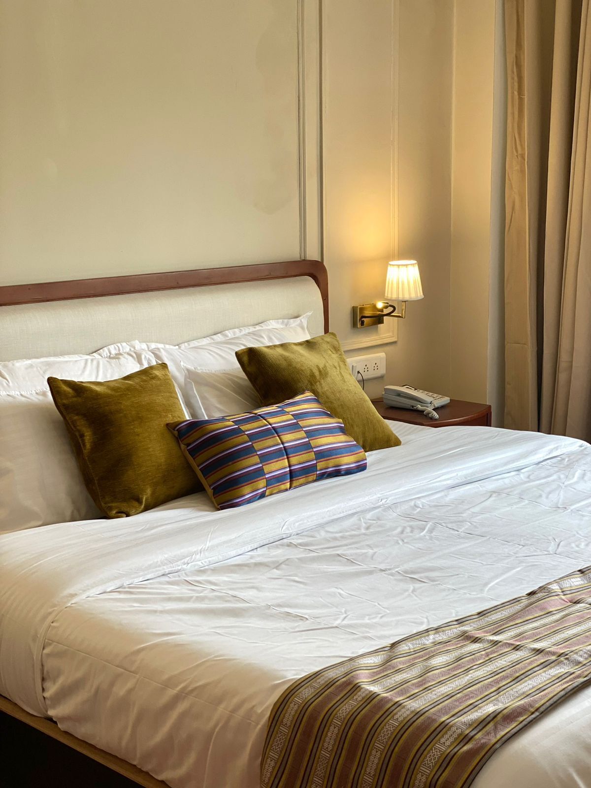 Ein Hotelzimmer mit einem ordentlich bezogenen Bett, einer Nachttischlampe, einem Telefon und einem Fenster mit Vorhängen.