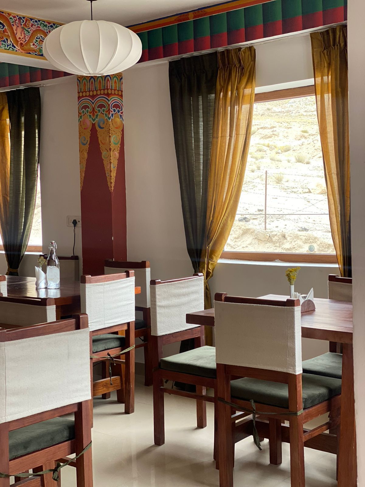 A dining area with wooden tables and chairs, a hanging light fixture, and a colorful wall decoration.