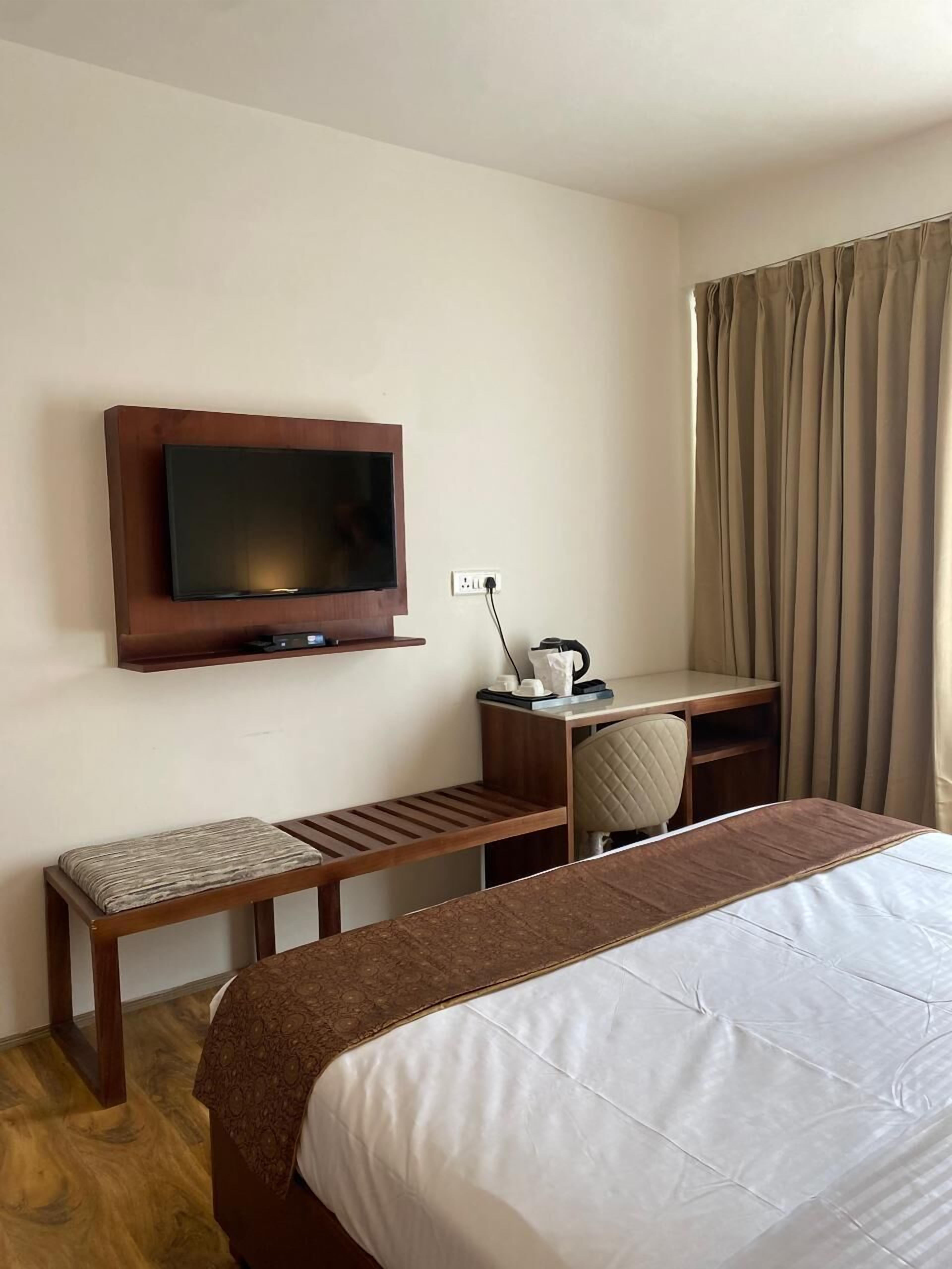 A hotel room with a bed, a TV mounted on the wall, a desk with a chair, and a window with curtains.