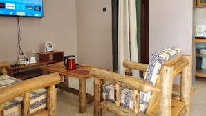 A living room with a wooden coffee table, a sofa with zebra-patterned cushions, a television mounted on the wall, and a kitchen area with a toaster.