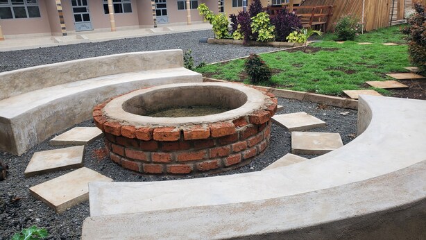 A circular brick fire pit with a concrete border, surrounded by stepping stones and gravel, set in a courtyard with a building in the background.