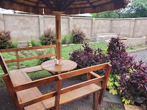 A wooden bench with a woven table in a garden area with various plants and a concrete wall in the background.