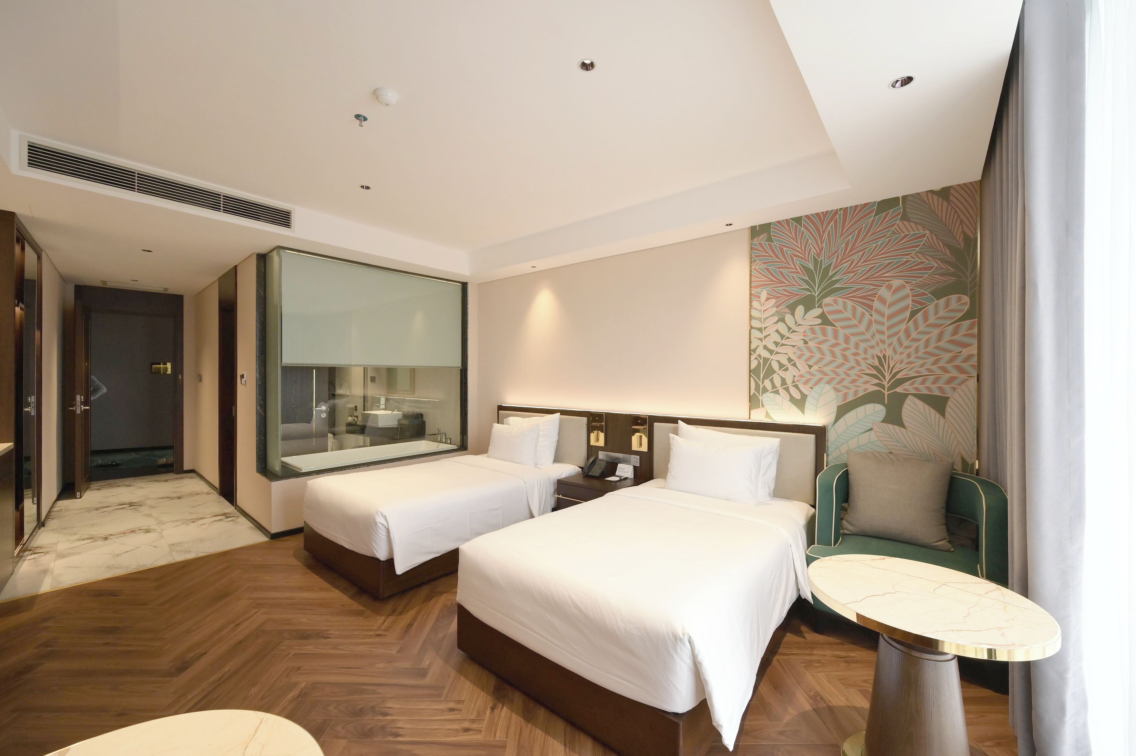 A hotel room with two beds, a sitting area, a small table, and a bathroom with a glass partition.