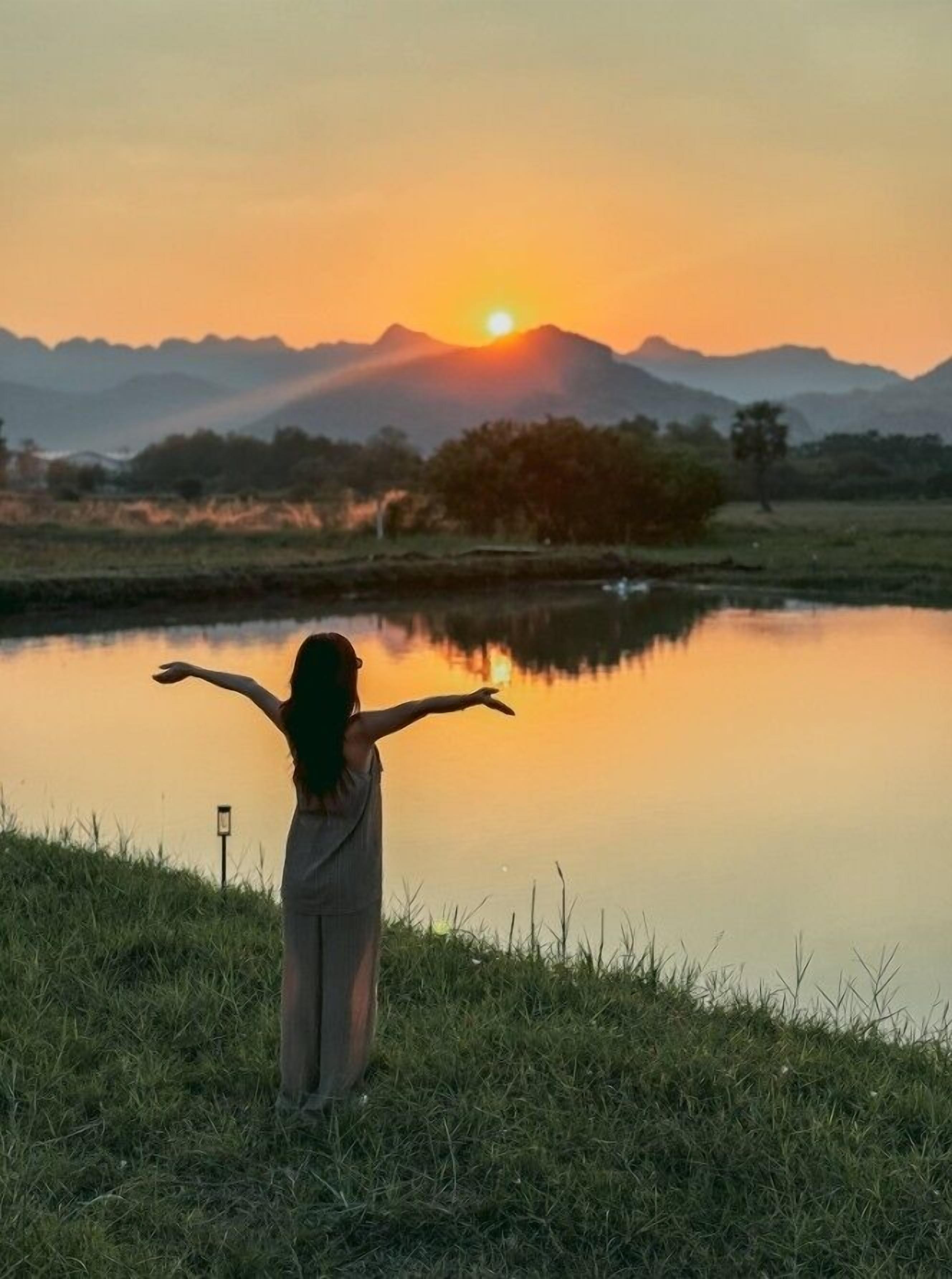 Une personne se tient debout, les bras tendus, face à un coucher de soleil sur une étendue d’eau, avec des montagnes en arrière-plan.