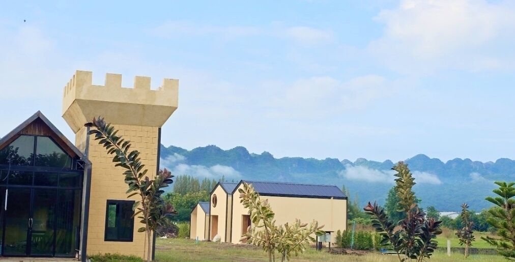 A castle-like structure with a tower, a modern building with a glass facade, and a small church-like building, all set against a mountainous backdrop.