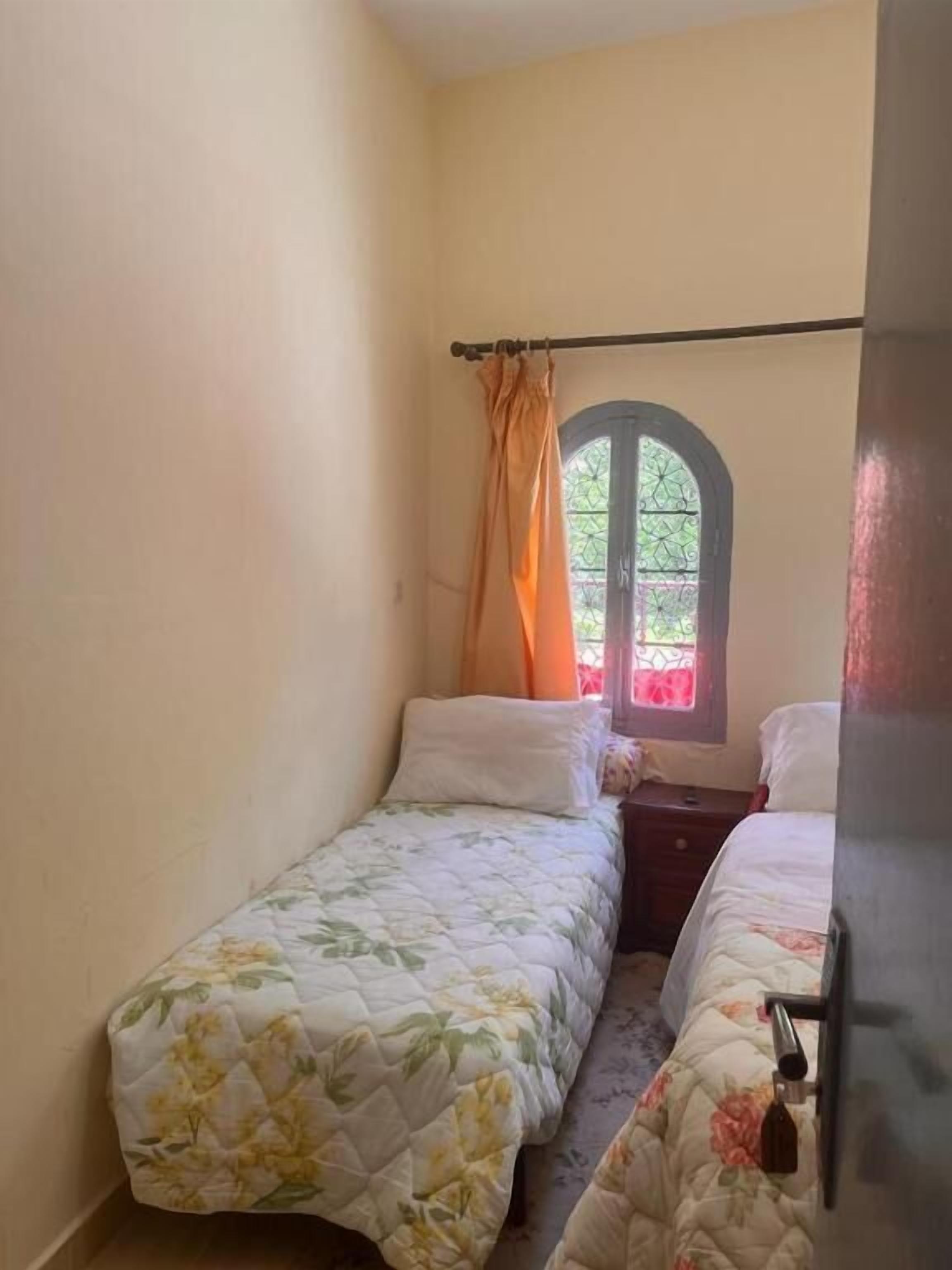 A small bedroom with two single beds, a window with a curtain, and a nightstand.