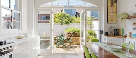 A bright kitchen with a large window, a dining area with a table and chairs, and a view of a garden through an open door.