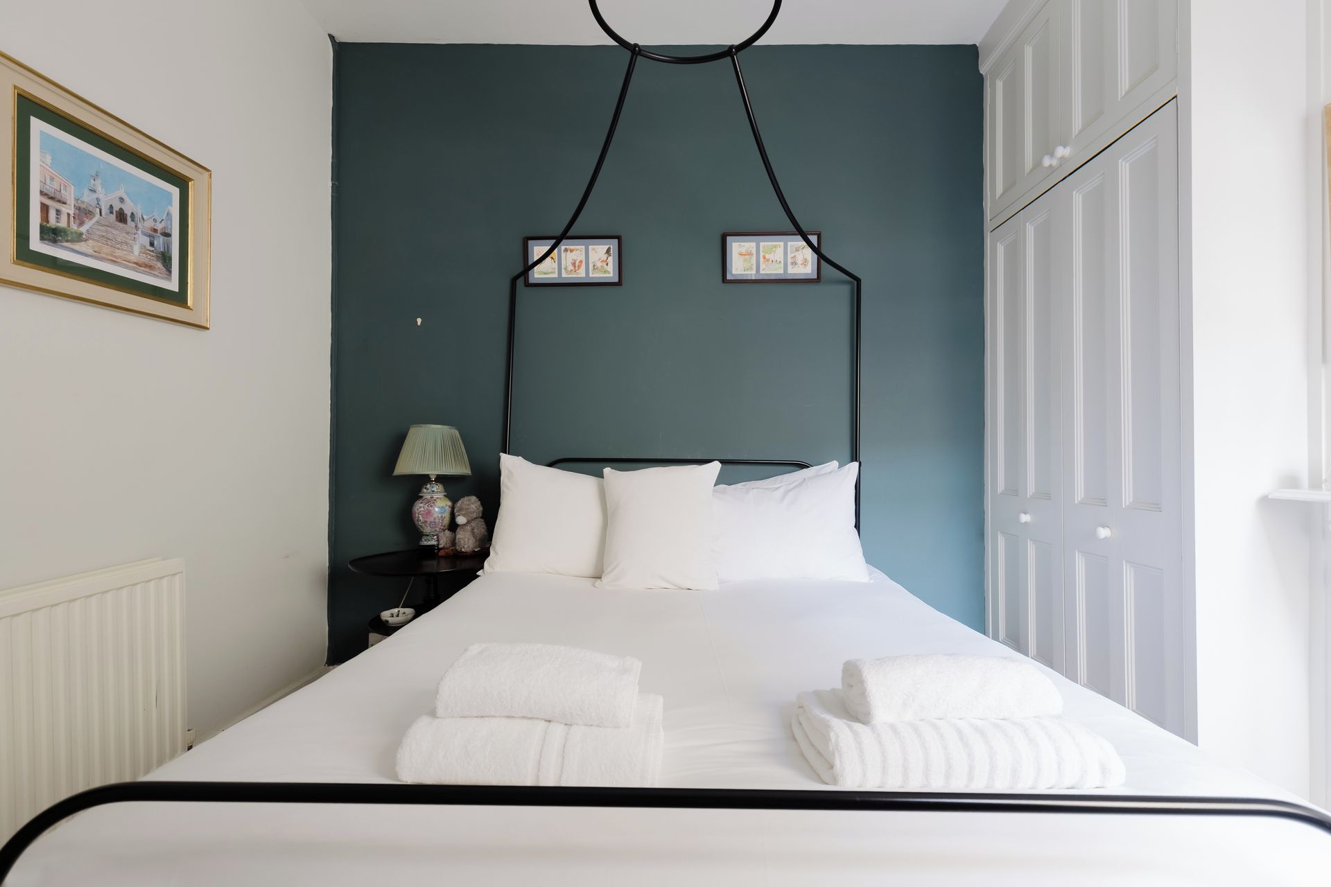 A neatly made bed with white linens and towels, a bedside lamp, a framed picture on the wall, and a door to the right.