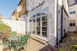 A white brick building with a large white-framed window, a satellite dish, and a small patio with a table and chairs. - Refined 2BD Home in Fulham (London)
