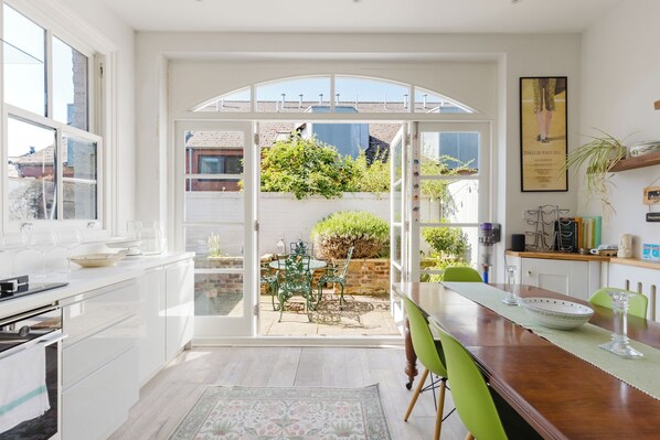 Eine helle Küche mit großem Fenster, einem Essbereich mit Tisch und Stühlen sowie einem Gartenblick durch eine offene Tür.