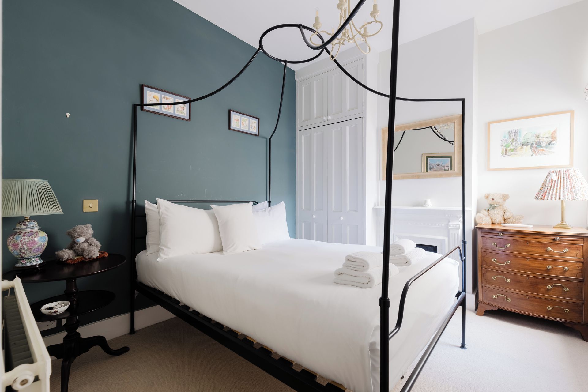 A four-poster bed with white linens, a bedside table with a lamp, and a dresser with drawers.