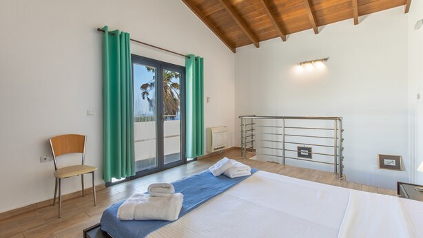 A bedroom with a bed, a chair, a balcony with curtains, and a view of palm trees.