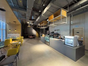 A modern cafe with yellow chairs, a counter with a display case, and a brick wall with 'THE HUB' signage. - Free housekeeping visits during your stay - StayShort - Stylish Studio in JVT Sleeps 2 with Balcony (JVT)