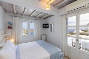 A bedroom with a bed, a TV, a window with curtains, and a view of the sea. - Mykonos Psarou Place, Seaview with Private Jacuzzi (Psarou)