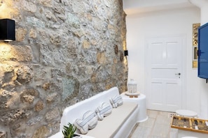 A bathroom with a stone wall, a white bathtub, a white door, and a blue cabinet. - Mykonos Psarou Place, Seaview with Private Jacuzzi (Psarou)