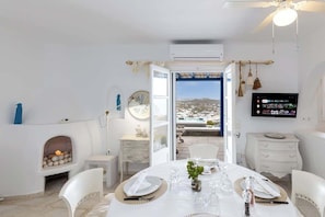 A room with a dining table set for four, a fireplace, a television, and a view of a coastal landscape. - Mykonos Psarou Place, Seaview with Private Jacuzzi (Psarou)