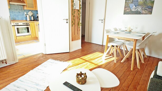 An open door leading to a kitchen with wooden cabinets and a tiled backsplash, a dining area with a white table and chairs, and a painting on the wall.