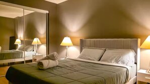 A neatly made bed with a green bedspread, two bedside lamps, a large mirror, and a nightstand with a lamp. - Main Downtown-Deluxe Suite w Pool, Gym & Balcony (Winnipeg)