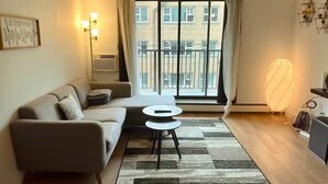 A modern living room with a sofa, a small round table, a standing fan, and a large window with curtains. - Main Downtown-Deluxe Suite w Pool, Gym & Balcony (Winnipeg)