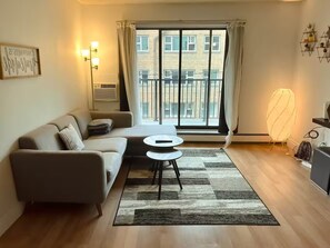 Een moderne woonkamer met een bank, een klein rond tafeltje, een vloerventilator en een groot raam met gordijnen.
