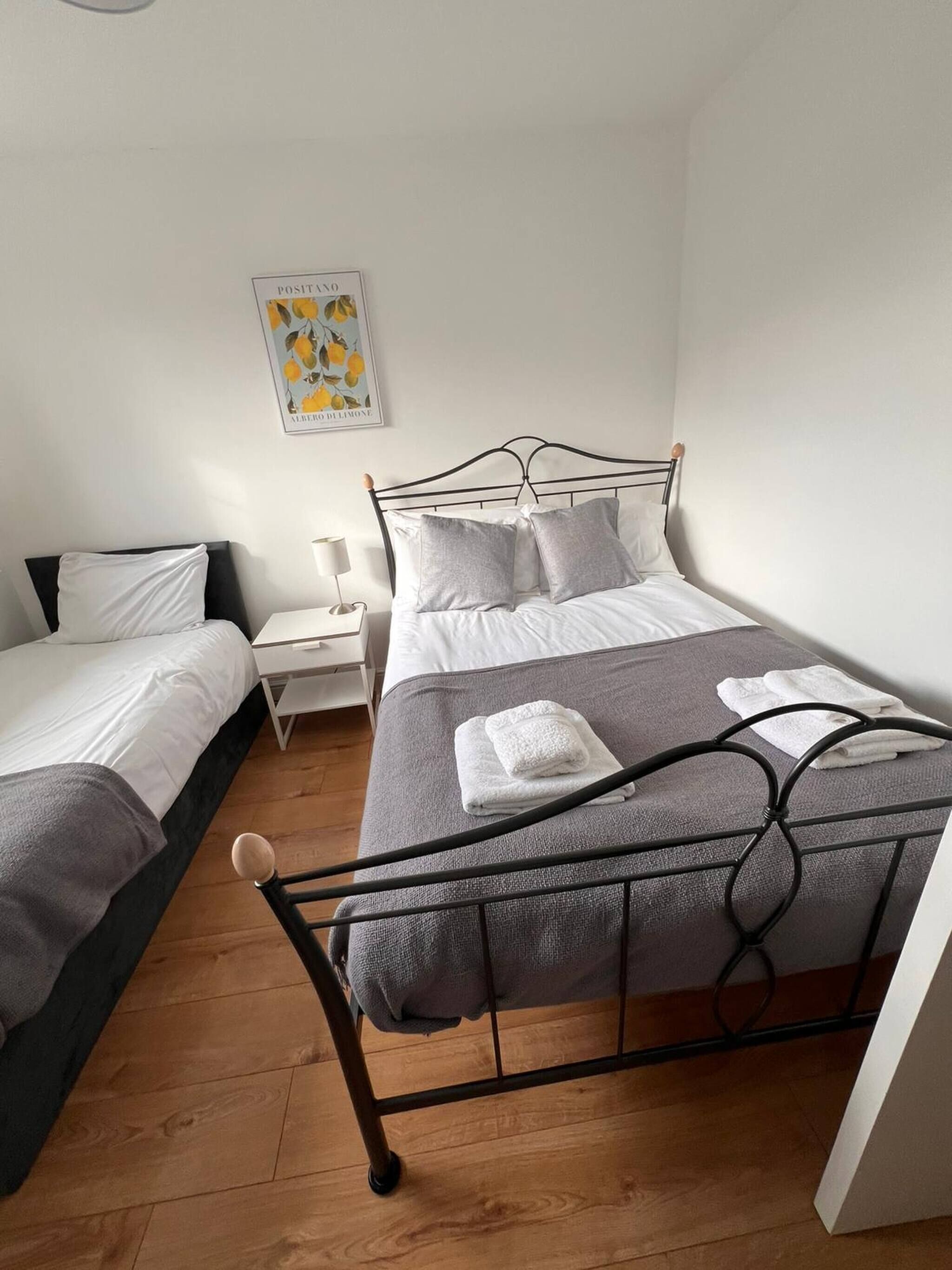 A bedroom with two beds, a bedside table with a lamp, a framed picture on the wall, and a wooden floor.