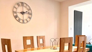 A dining area with a wooden table and chairs, a wall clock, and a vertical panel. - Tranquil Turquoise - 3 Bed House With Parking (Bracknell)