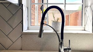 A kitchen with a double stainless steel sink, a modern faucet, and a window with a view of a brick building and a wooden fence. - Tranquil Turquoise - 3 Bed House With Parking (Bracknell)
