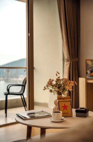 Una mesa con una taza de café, un libro, un florero, una libreta y una pequeña caja, con una silla y una ventana al fondo.