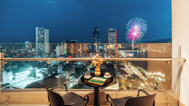 Un balcone con due sedie, un tavolo con un libro e un vaso, e sullo sfondo un panorama urbano con edifici e fuochi d'artificio.