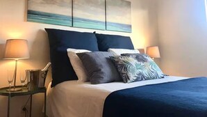 Ein Hotelzimmer mit Bett, zwei Nachttischlampen, einem Nachttisch mit Lampe und einem Bild von einer Strandlandschaft.