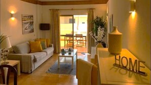 Ein Wohnzimmer mit Sofa, Couchtisch und Fernseher. Es gibt eine Essecke mit Tisch und Stühlen. Der Raum strahlt eine warme Atmosphäre aus, ergänzt durch an der Wand befestigte Lampen.