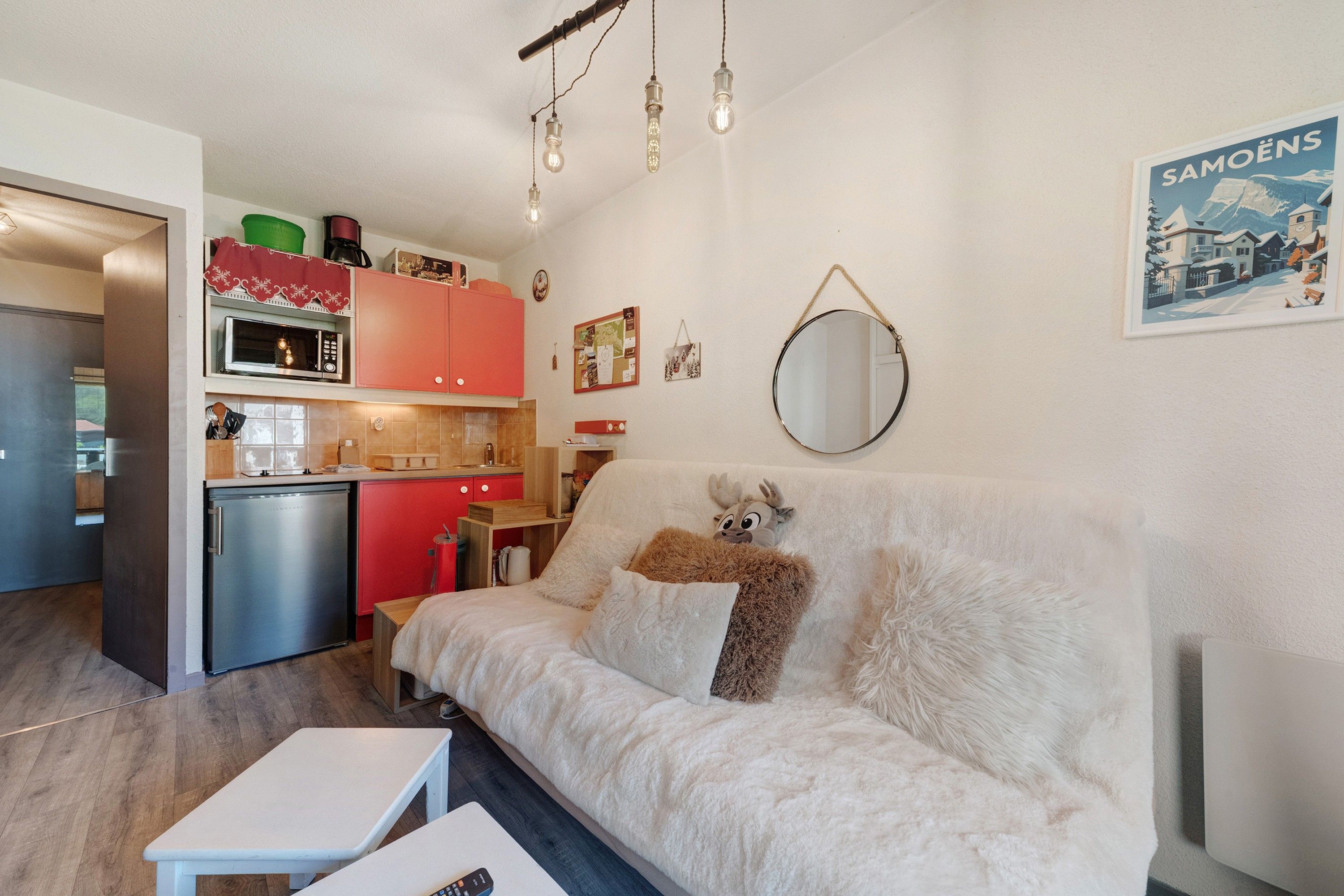 A cozy living room with a white sofa, a small table, and a kitchen area with red cabinets and a microwave.