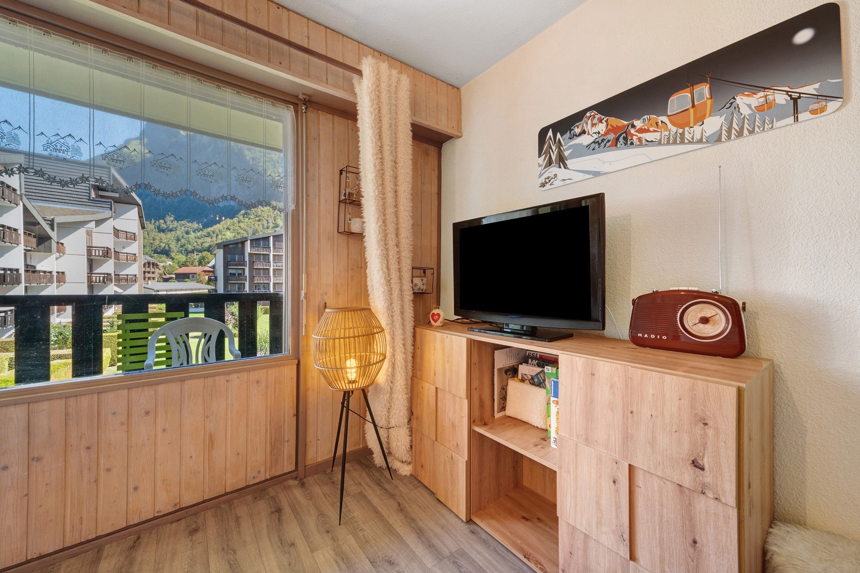 Ein Zimmer mit großem Fenster, Fernseher, Radio und einem Bücherregal.