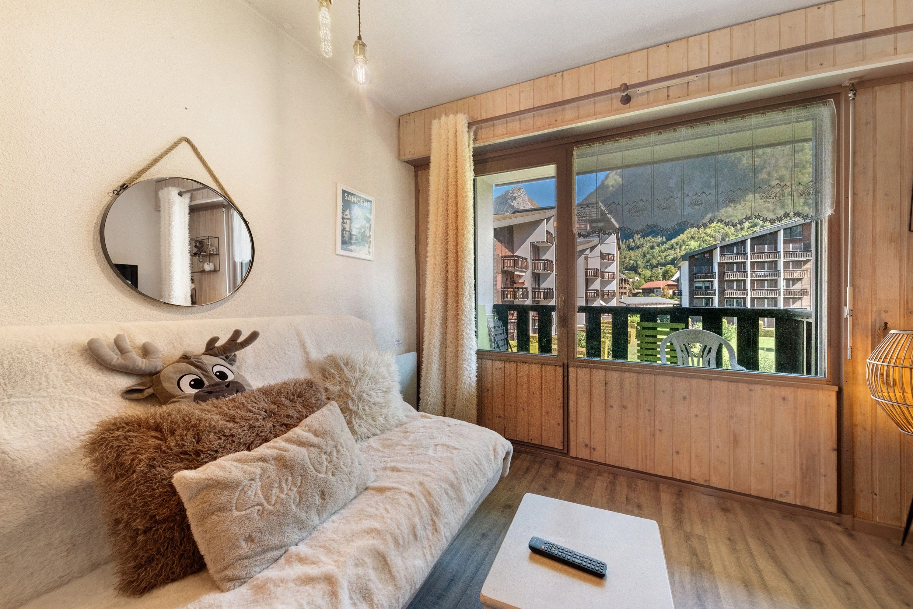 Una habitación acogedora con piso de madera, un sofá con un peluche de reno, un espejo redondo y una ventana con vistas a edificios.