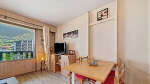 Ein Zimmer mit Holzboden, einem Esstisch mit Stühlen, einem Fernseher und einem Fenster mit Blick auf Gebäude und Grünflächen.