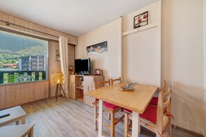 Une pièce dotée d’un plancher en bois, d’une table à manger avec des chaises, d’un téléviseur et d’une fenêtre donnant sur des bâtiments et de la verdure.